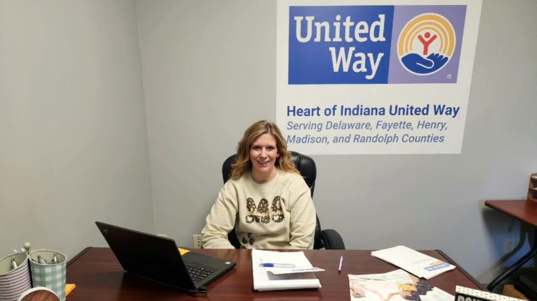 Ashley sits behind a desk in Heart of Indiana United Way's new Randolph County office. The desk has a laptop and papers. On the back wall there is a large United Way logo.