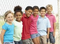 6 Diverse Children with their arms around each other stand against a playground fence.